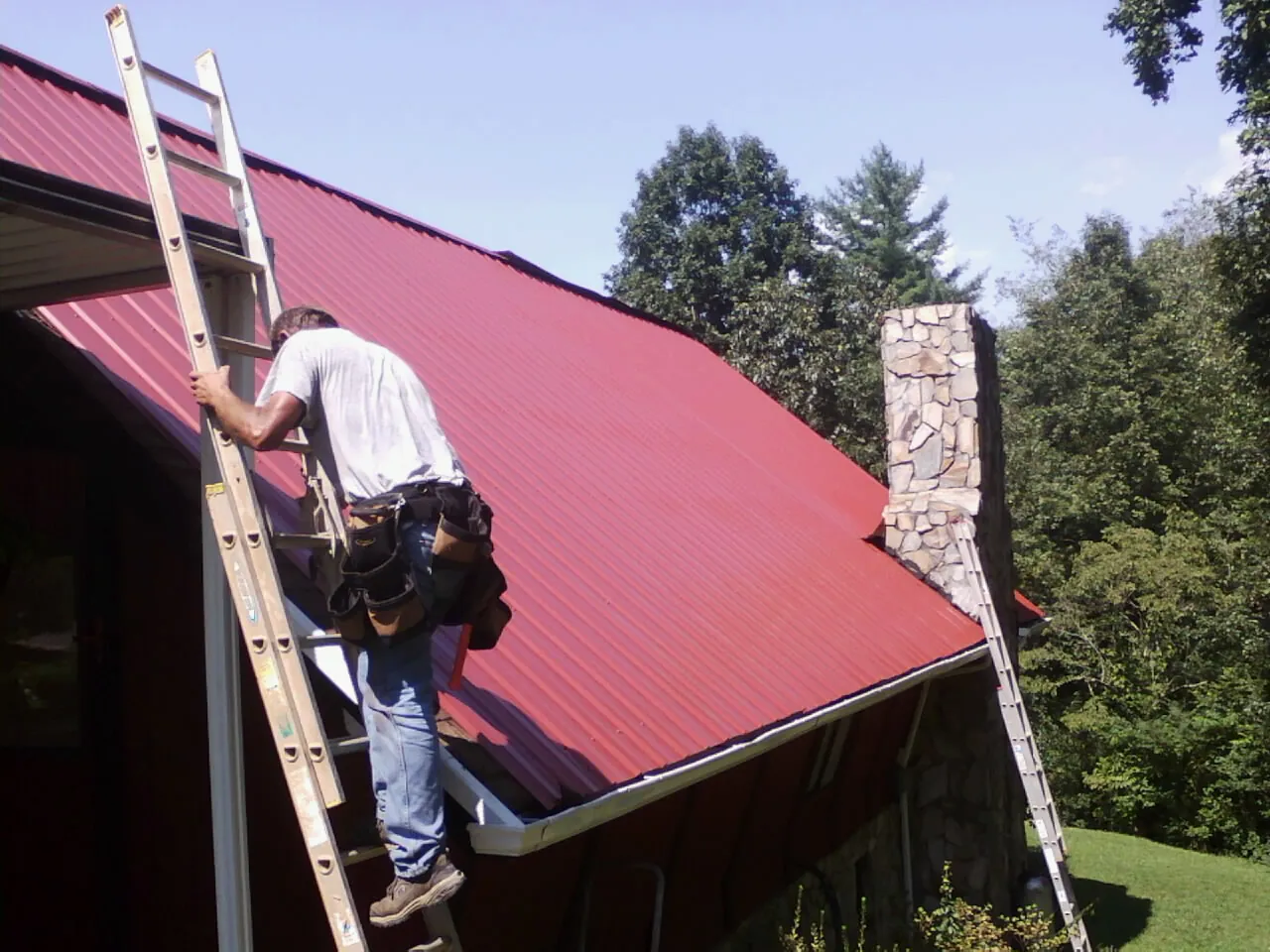 Roofer on a ladder beside a red metal roof and stone chimney — Soffit & Fascia Repair by Roofing Brothers in Loami